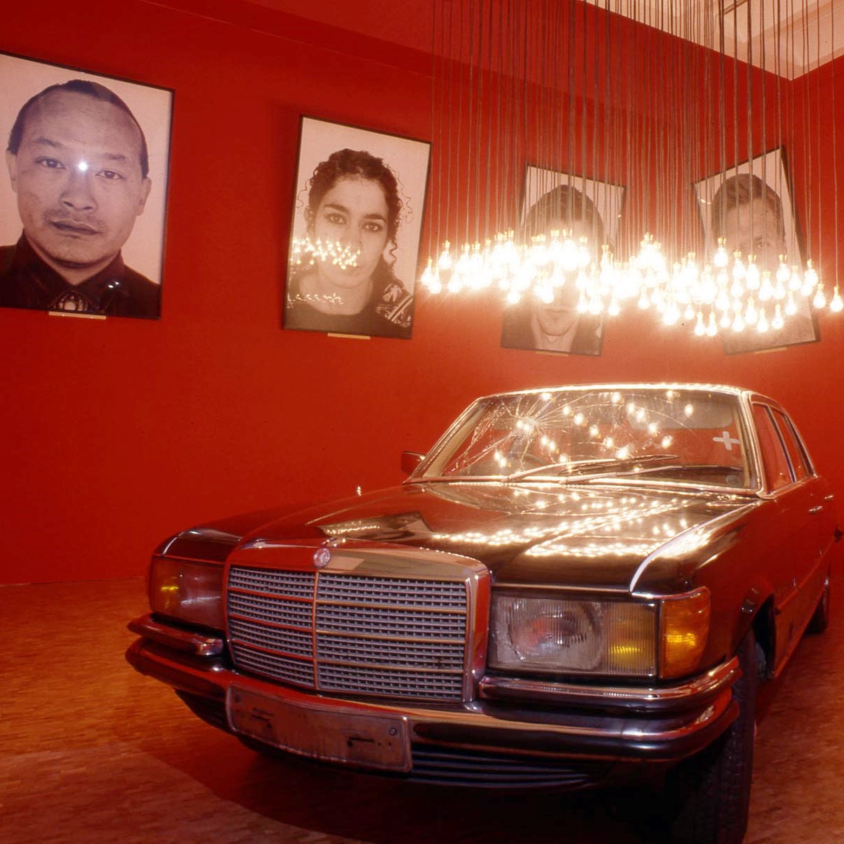 Salle d'exposition rouge avec portraits photographiques aux murs et une voiture sous un luminaire au centre.