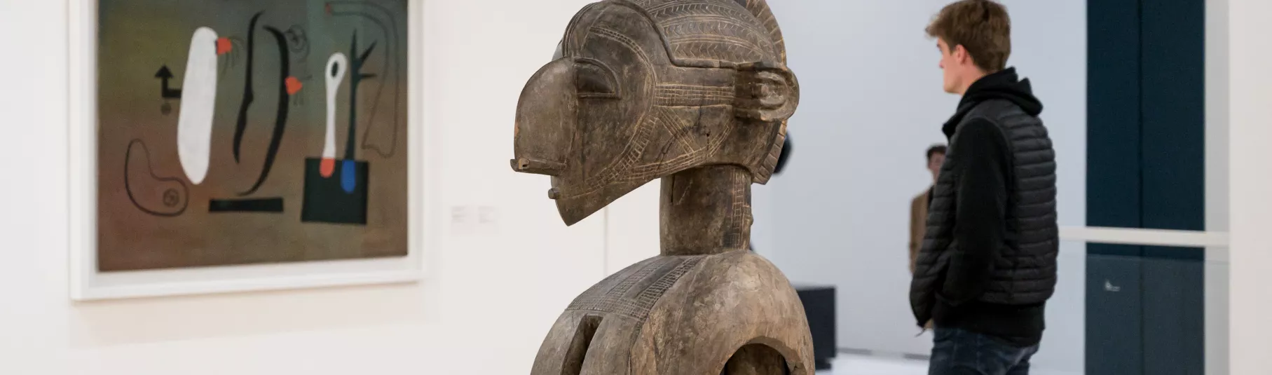 Salle d'exposition du musée avec une sculpture au centre, un tableau et un visiteur.
